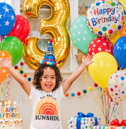 Happy child celebrating a third birthday surrounded by colorful helium balloons, a giant gold number 3 balloon and Happy Birthday balloons.